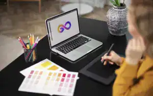  A graphic designer uses a stylus on a drawing tablet, creating a logo on a laptop screen. Color swatches and pencils are on 