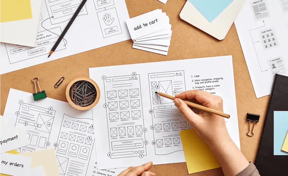A top-down view of a person sketching a website layout on a desk. The desk is covered with various wireframe papers, notes, p